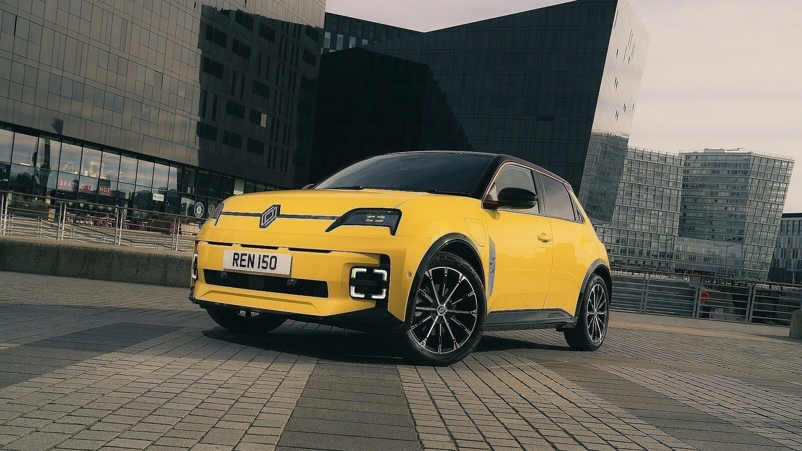 Şehir mimarisi önünde sarı Renault 5 E-Tech Elektrikli ön üç çeyrek görünümü